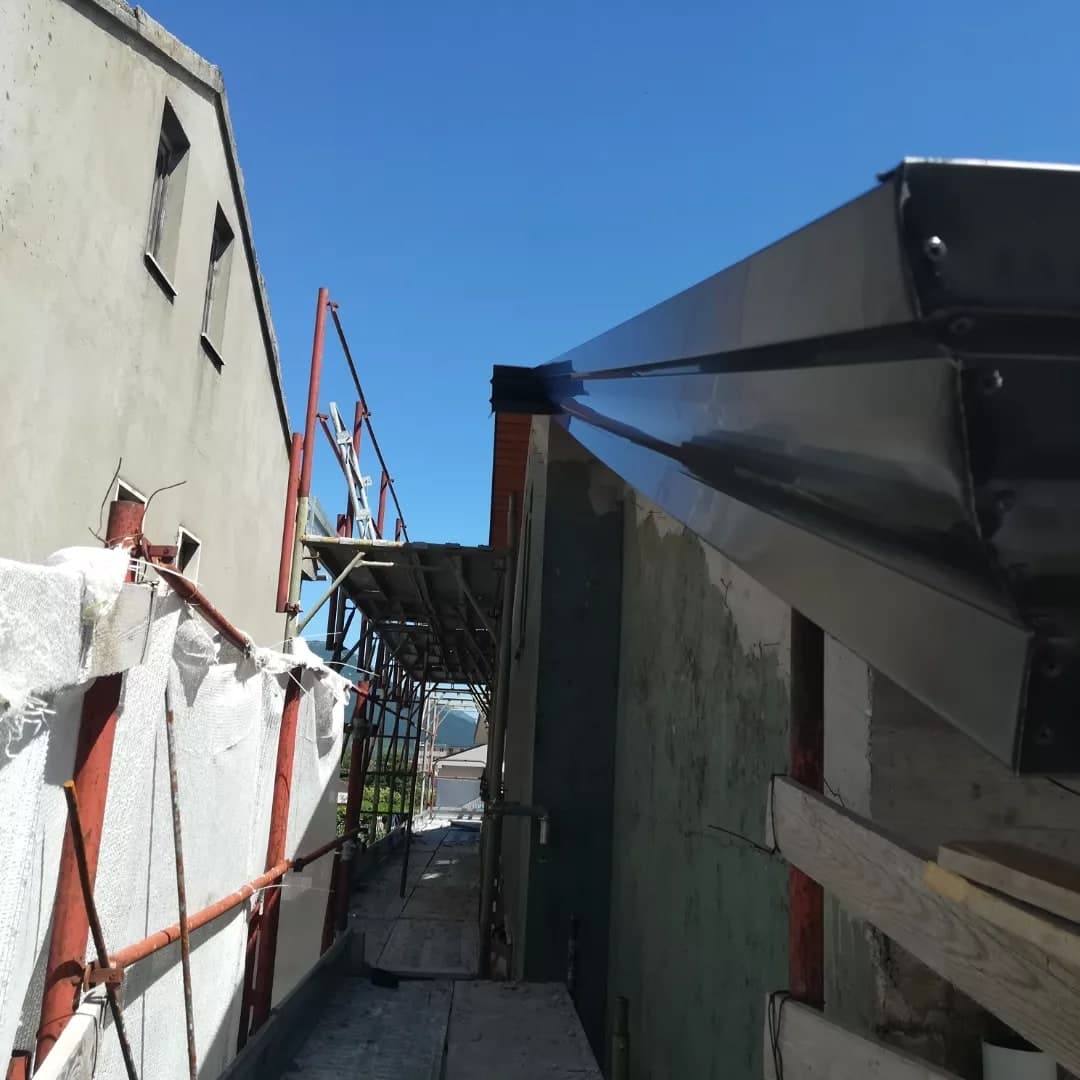 Dalla strada stretta: la gronda scura all’angolo della casa vista da sotto, con la testata; accanto, il ponteggio.