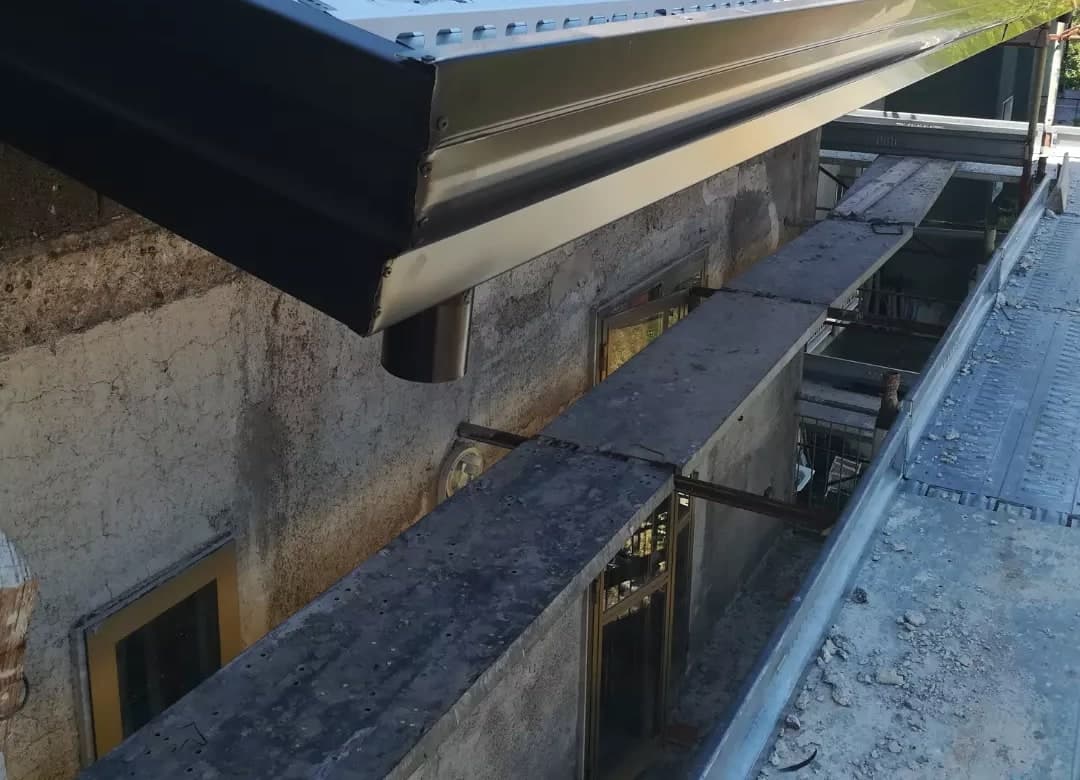Dal ponteggio: la gronda scura a gradini lungo il bordo del tetto, con il bocchello del pluviale sotto; sopra, la lamiera grecata grigia che finisce dentro il canale.