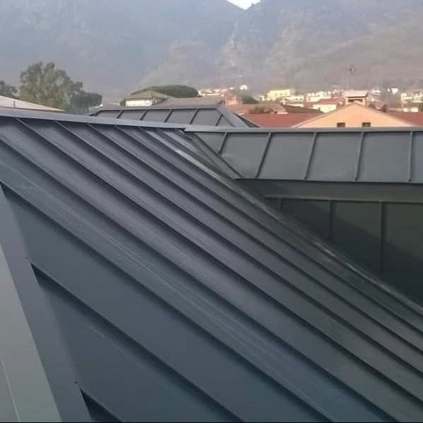 Le falde in lamiera aggraffata antracite, con le pieghe dritte; dietro, i tetti di un paese di valle e i monti.