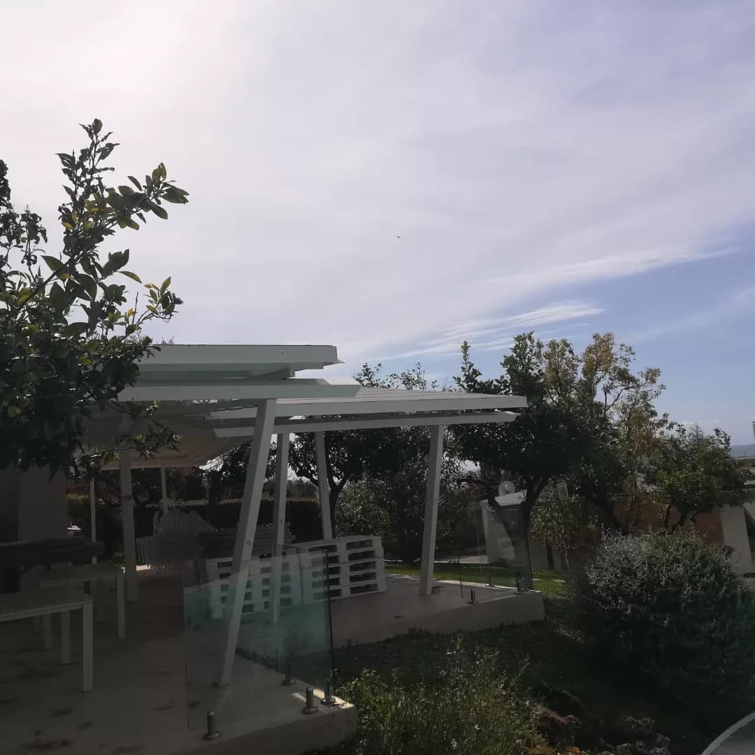 La pergola bianca vista dal giardino, con i montanti inclinati, sulla terrazza con il parapetto di vetro.