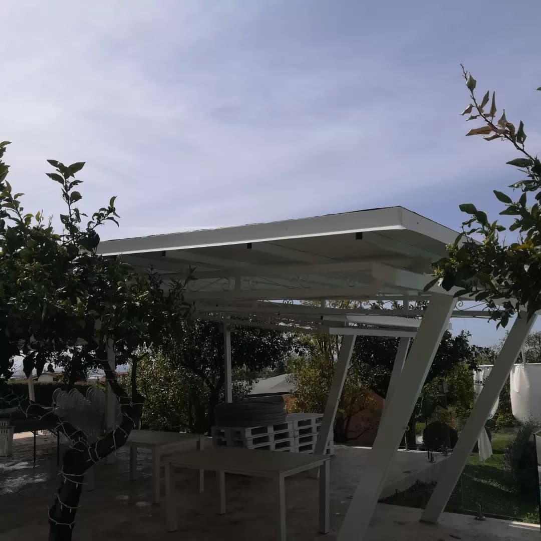 La pergola bianca fra gli agrumi: la copertura piana con il bordo bianco alto, sotto le travi bianche con un fregio di ferro battuto, e i montanti inclinati tutti dalla stessa parte; sotto, tavoli e bancali.