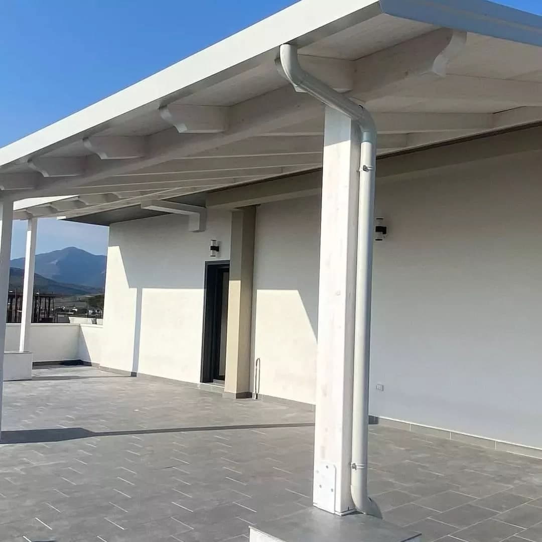Sul terrazzo dell’attico, la tettoia in legno bianco: il pluviale bianco fa il collo d’oca dalla gronda e scende lungo il pilastro con due collari, fino al plinto; sotto il bordo, le teste sagomate dei travetti; dietro, il muro dell’attico con la porta.