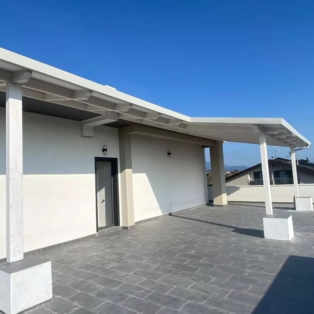 La tettoia bianca lungo il muro dell’attico e, oltre il suo spigolo, il pezzo più basso su altri pilastri; i pilastri stanno sui plinti di calcestruzzo, il terrazzo è in piastrelle grigie con il parapetto bianco.