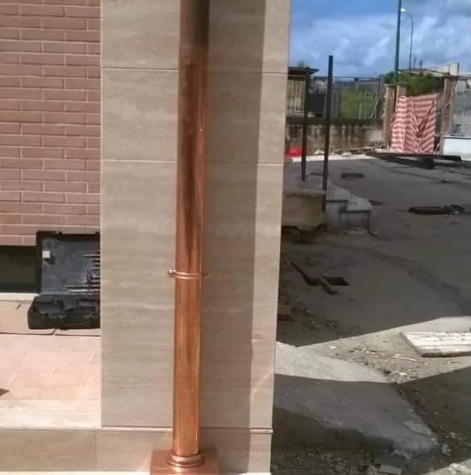 Lo stesso pilastro da un passo più in là: il pluviale di rame lungo la pietra, il collare, la cassetta al piede; dietro, il cortile del cantiere.