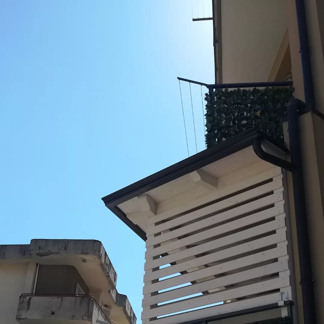 La tettoia dal fianco sinistro, da sotto: la gronda scura lungo il bordo e, dal suo capo contro la facciata, il pluviale con il collo d’oca che scende lungo il muro; sotto, le doghe bianche del ripostiglio.