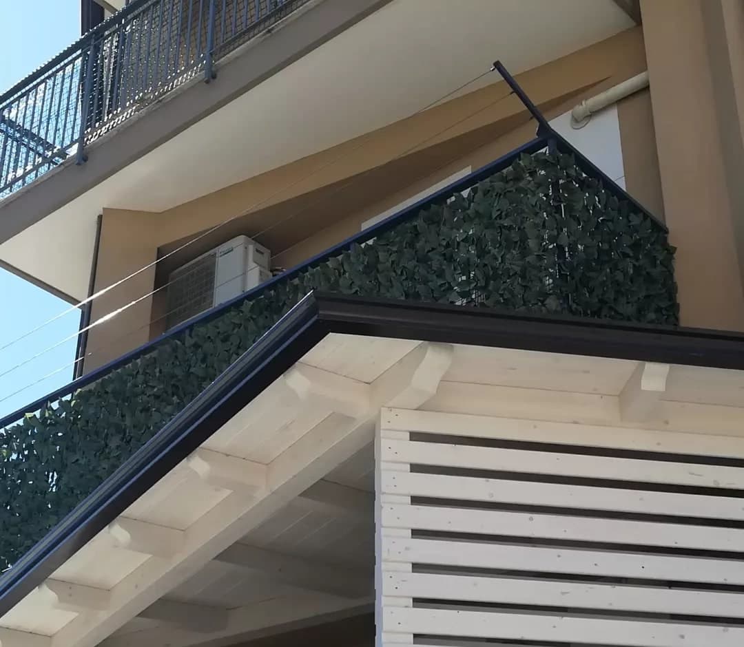 La tettoia di legno bianco sul terrazzo, da sotto: la gronda testa di moro corre sul fianco e gira l’angolo sul davanti; sotto, i travetti con le teste sagomate e il ripostiglio a doghe orizzontali; sopra, la ringhiera del balcone coperta di finta edera.