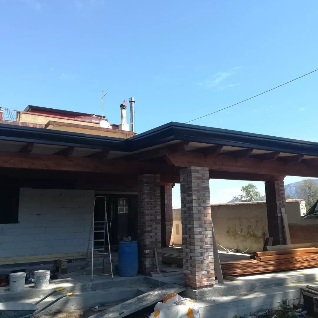 Il portico in legno davanti a una casa a un piano ancora in costruzione: pilastri in mattoni, travi a vista e la gronda scura che corre lungo il bordo.