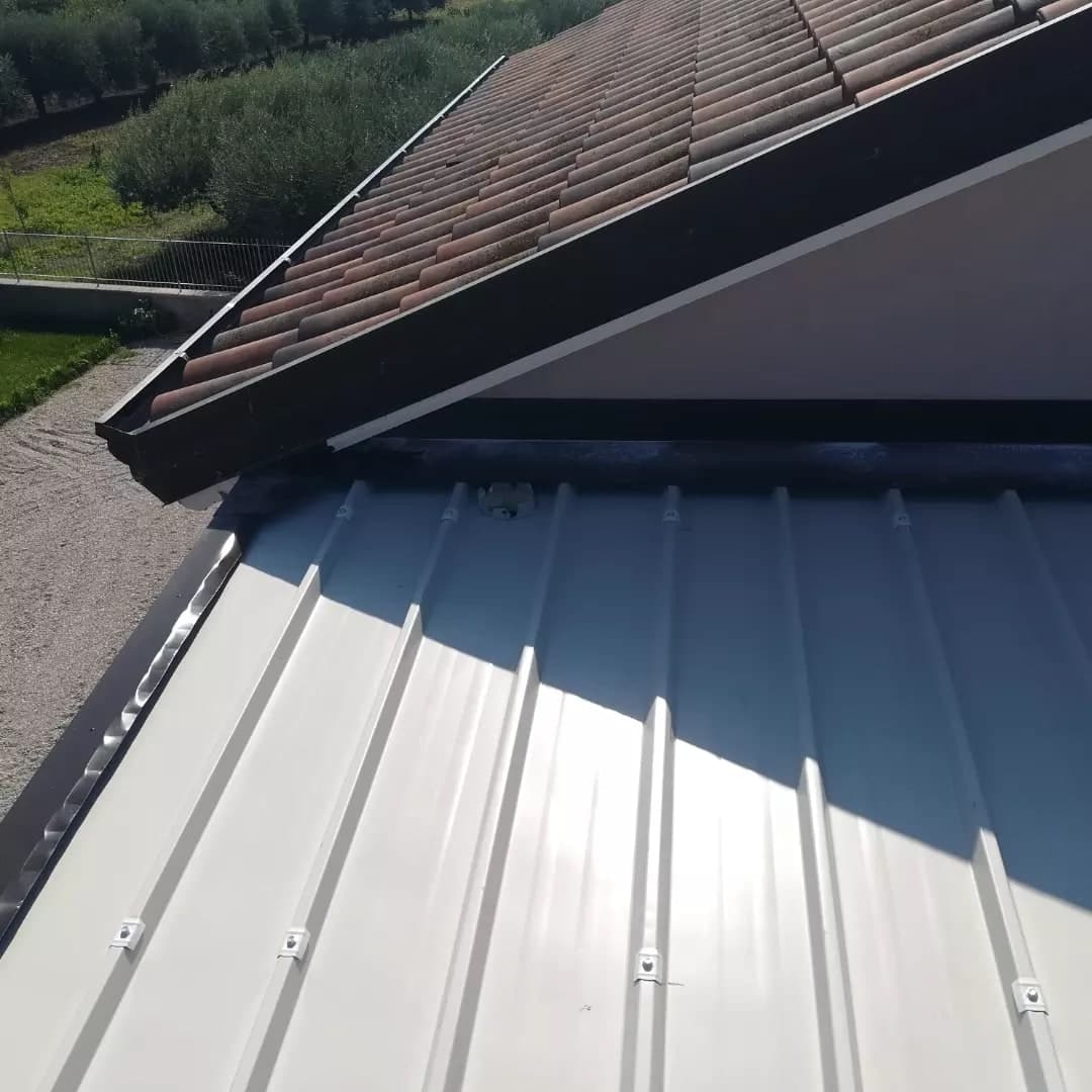 Dall’alto, la lamiera grecata grigio chiaro della tettoia con le viti sulle creste, la scossalina scura sul fianco e contro il timpano; sopra, il timpano della casa con i coppi e la scossalina scura sugli spioventi.
