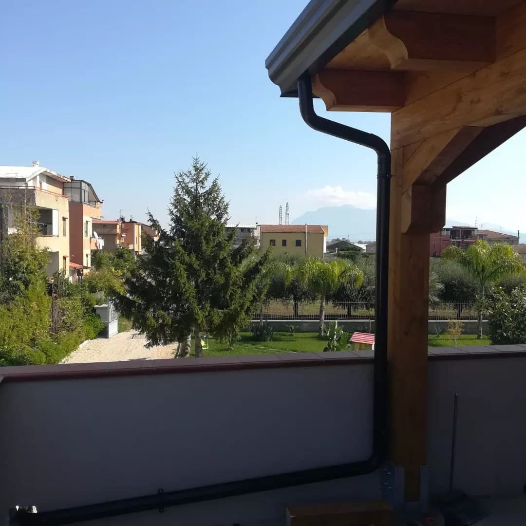 Dal terrazzo, il pilastro d’angolo della tettoia: la gronda a cornice antracite con le teste sagomate dei travetti, il pluviale scuro con il collo d’oca lungo, giù lungo il pilastro e poi steso sul pavimento lungo il parapetto; oltre il parapetto, il giardino, le palazzine e i monti.