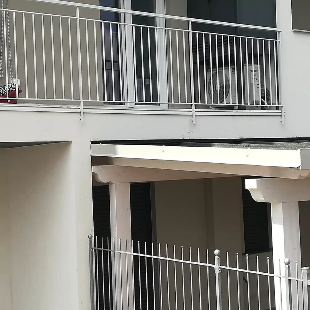 Sotto un balcone, il bordo della tettoia: la lamiera bianca con le teste delle viti, sopra la guaina grigia; sotto, le travi bianche con il capo sagomato sui pilastri.
