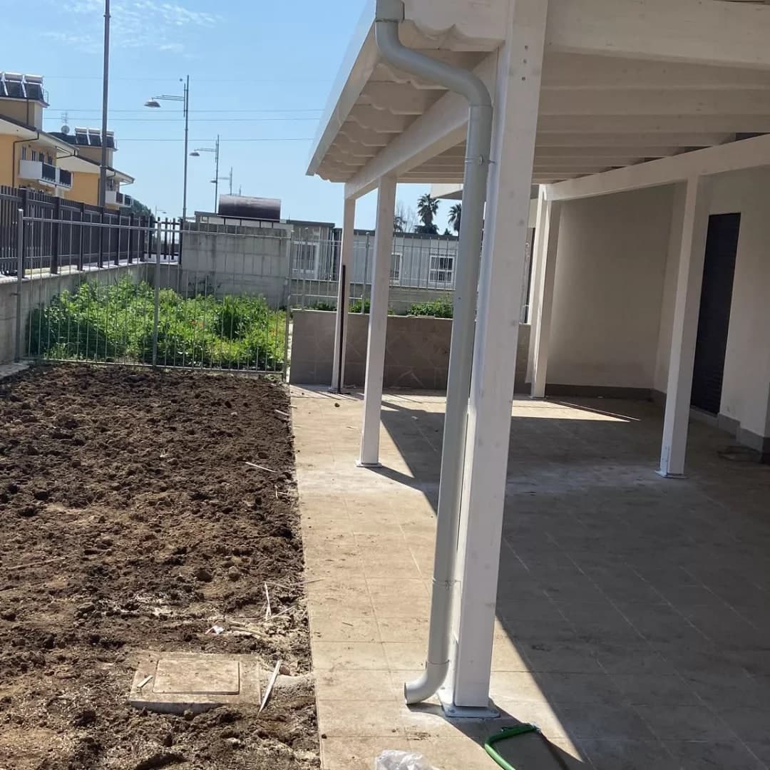 Sotto la tettoia, il pluviale bianco: dalla gronda fa il collo d’oca, scende lungo il pilastro d’angolo con i collari e al piede gira verso il giardino; sopra, i travetti bianchi con le teste sagomate; a destra il muro della palazzina, a sinistra l’aiuola appena vangata.