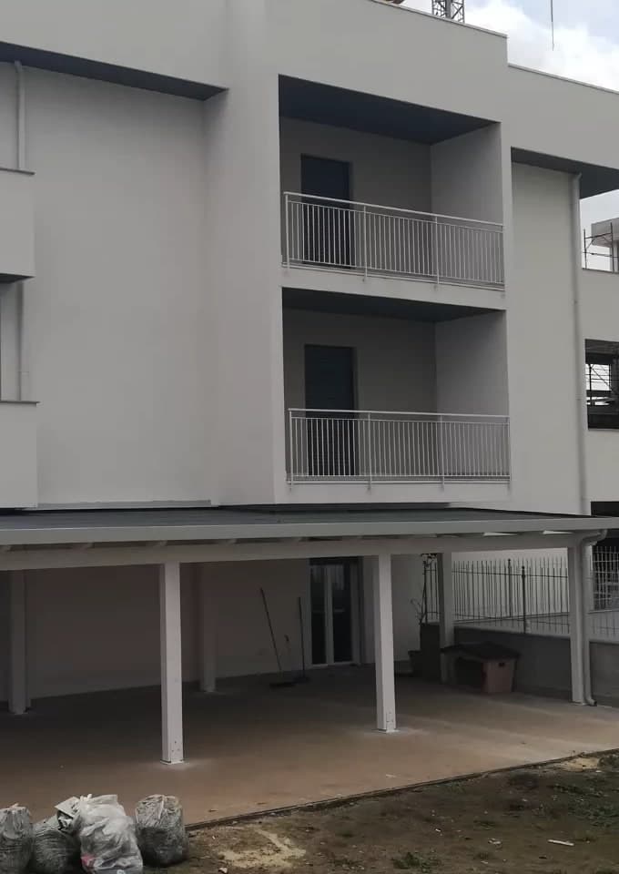 La palazzina bianca dal giardino e, lungo il piano terra, la tettoia in legno bianco sotto i balconi del primo piano: i pilastri sul bordo libero, la trave, il bordo bianco; a destra il pluviale bianco sul pilastro d’angolo.