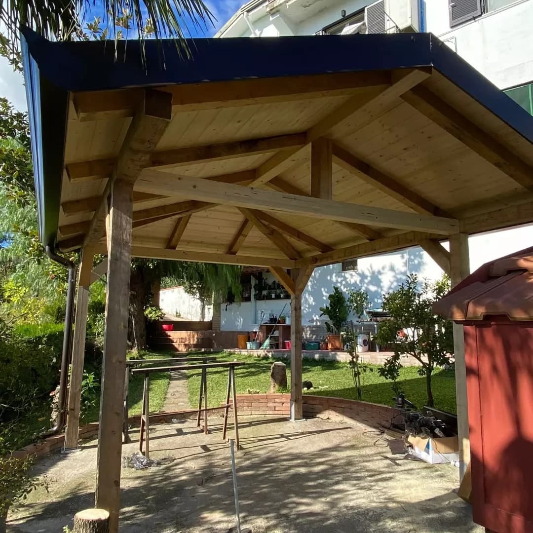 Il gazebo da sotto: i pilastri con le saette, le travi, i puntoni di displuvio e il monaco sulla catena, i travetti e il tavolato di abete chiaro; la gronda nera lucida riflette il cielo; a sinistra il pluviale; dietro, il giardino e una casa bianca.