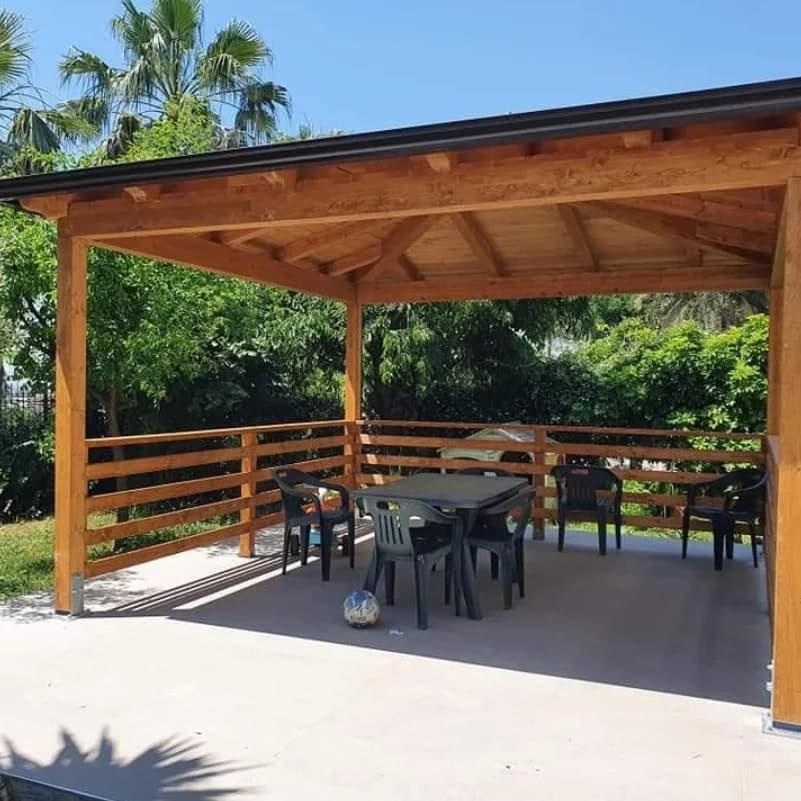 Il gazebo in legno color miele, finito, sulla platea di cemento: lungo il bordo del tetto la gronda scura, sotto i travetti e i puntoni a vista; il parapetto di tre listoni sui lati, dentro un tavolo e le sedie da giardino.