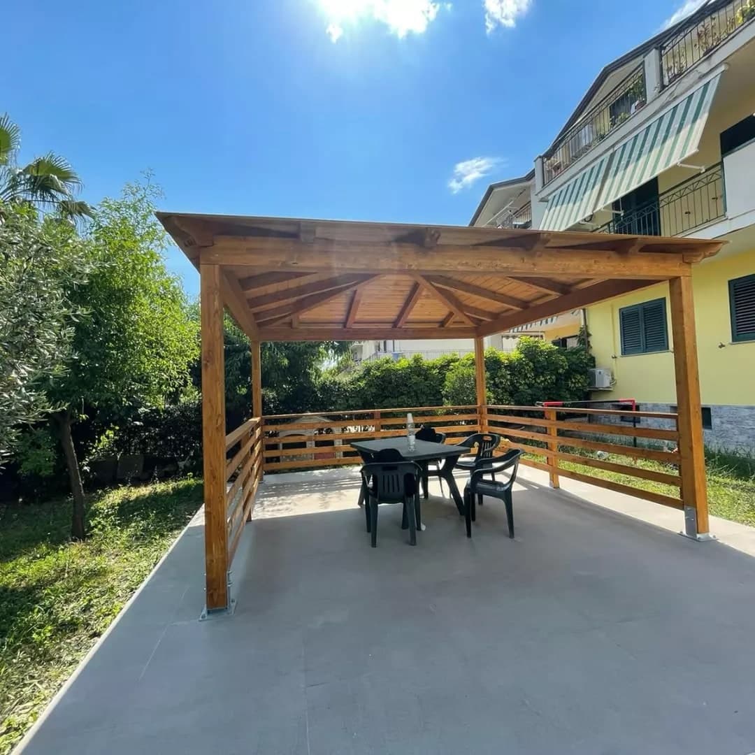 Il gazebo dal davanti aperto, da sotto: i travetti, i puntoni che salgono alla punta, il parapetto; a destra la palazzina gialla con i balconi e le tende.