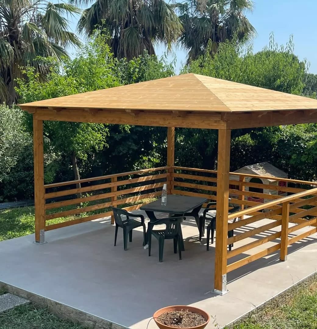 Il gazebo dall’alto, prima del manto: il tetto a punta in tavolato chiaro, i quattro pilastri sulle scarpe d’acciaio, il parapetto di tre listoni su tre lati e il davanti aperto; attorno il giardino con le palme.