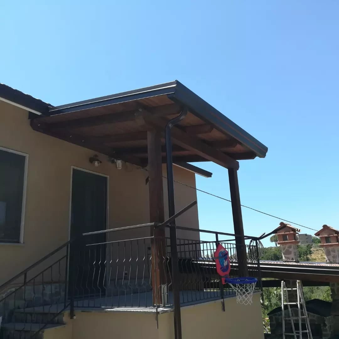 Dal basso, il portico d’ingresso in legno scuro sul pianerottolo della scala, con la gronda e il pluviale scuri; a destra, oltre la ringhiera, il tetto del porticato con i due comignoli, sui pilastri di pietra.