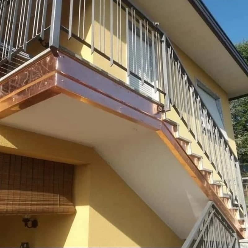 Il pianerottolo e la scala di una casa gialla, da sotto: la fascia di rame nuovo gira l’angolo del pianerottolo, a due fasce, con le viti; lungo la scala il rame segue i gradini a dente di sega e il bordo inclinato della soletta. Sopra, la ringhiera di piatti chiari; sotto il pianerottolo una tenda di bambù.