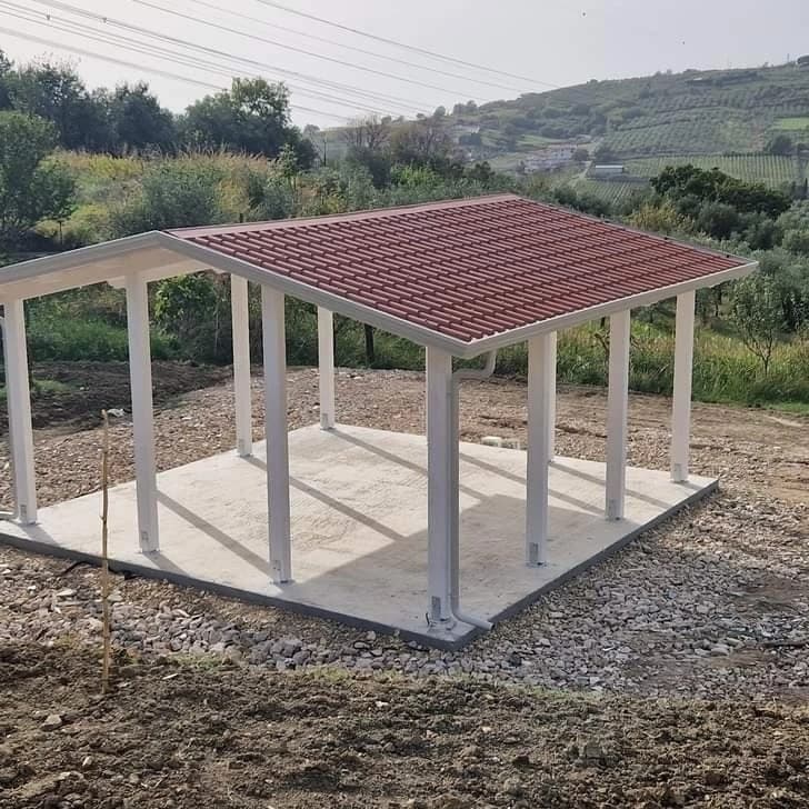 Una tettoia in legno bianco su una platea di cemento, vista dall’alto: il manto rosso a file di coppi, la gronda lungo il bordo e il pluviale che scende sul pilastro d’angolo.