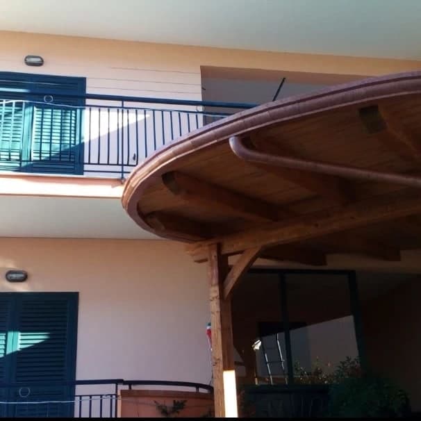 La pensilina curva vista da sotto: il tavolato e i travetti a raggiera in legno chiaro, il pilastro con le saette, lungo il bordo la gronda curva in rame e il pluviale che parte sotto.