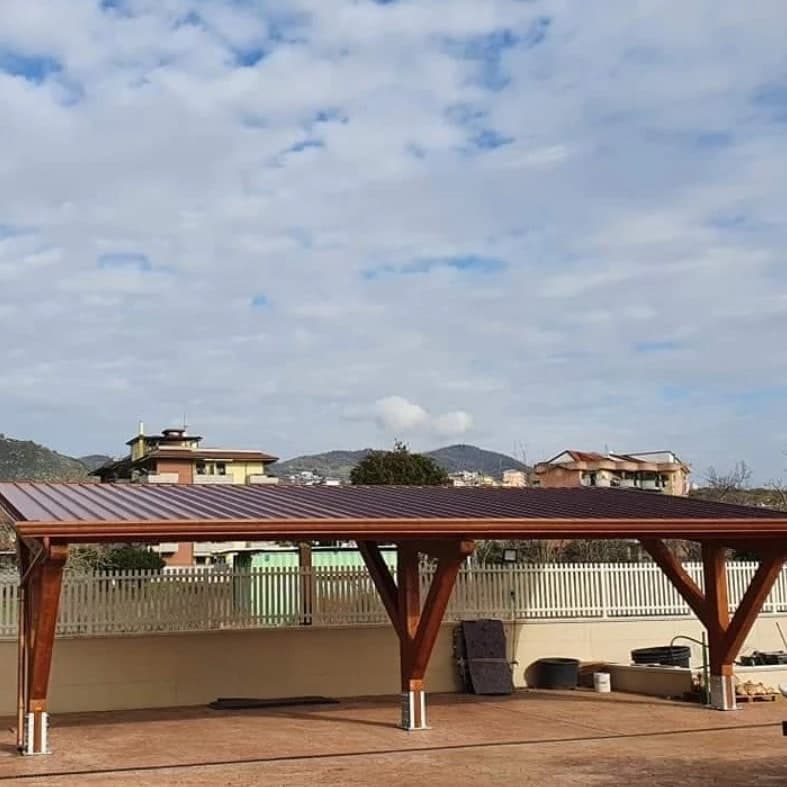 Una tettoia in legno lamellare vista dal lato lungo: la falda bassa in pannelli a nervature color marrone rame, la gronda color rame lungo il bordo, i pilastri che si aprono a Y sotto le travi.