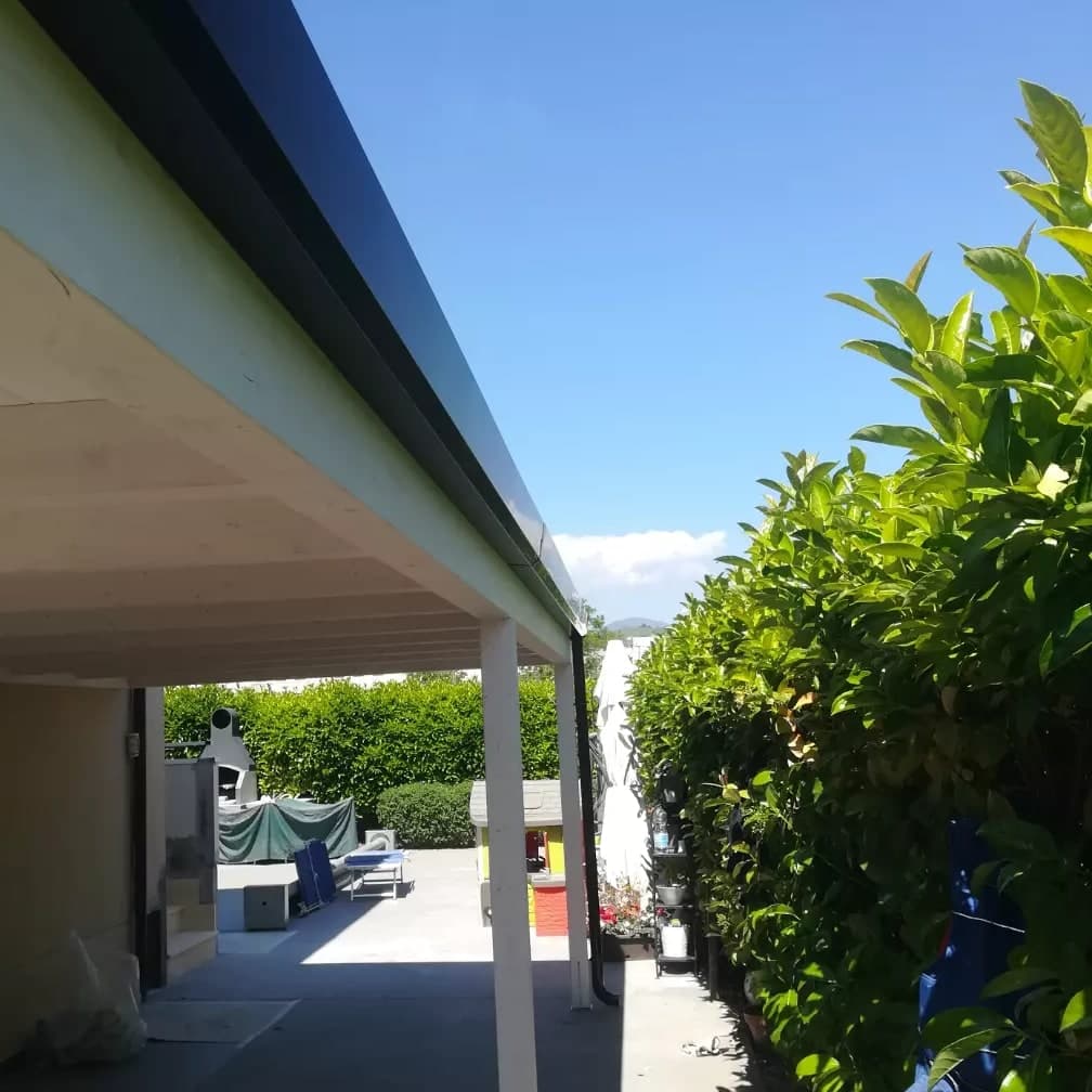 Da sotto la tettoia in legno bianco, lungo il fianco della casa: la gronda nera lucida corre sulla trave fino all’angolo in fondo, dove il pluviale nero scende accanto al pilastro e finisce con la curva al piede; a destra la siepe, in fondo il giardino.