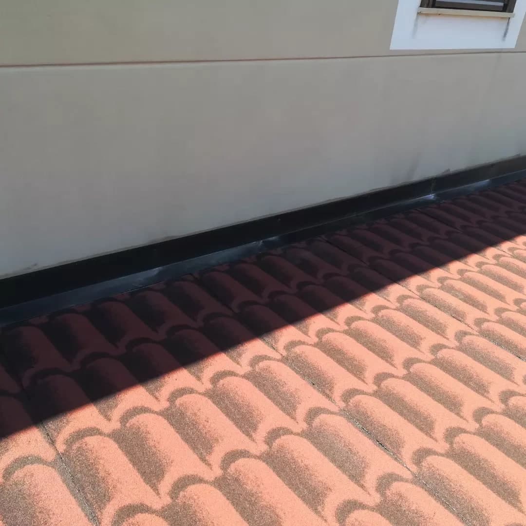 La guaina stampata a coppi rossi contro il muro chiaro della casa, con la scossalina nera che risale sul muro; sopra, la fascia e una finestra dalla cornice bianca.
