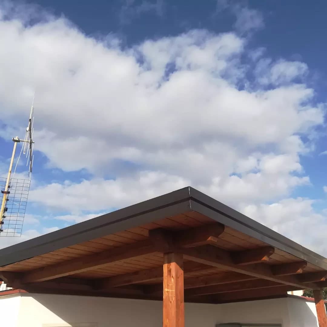 La tettoia contro il cielo, dall’angolo: la stessa faccia scura sul davanti e sul fianco, la trave e i travetti, due pilastri; dietro, il muro bianco del terrazzo.
