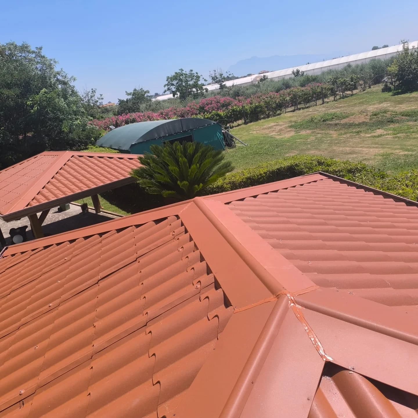 Dall’alto, i tetti rossi di due gazebo nel giardino: il colmo e i displuvi coperti di lamiera rossa; sullo sfondo campi e serre.