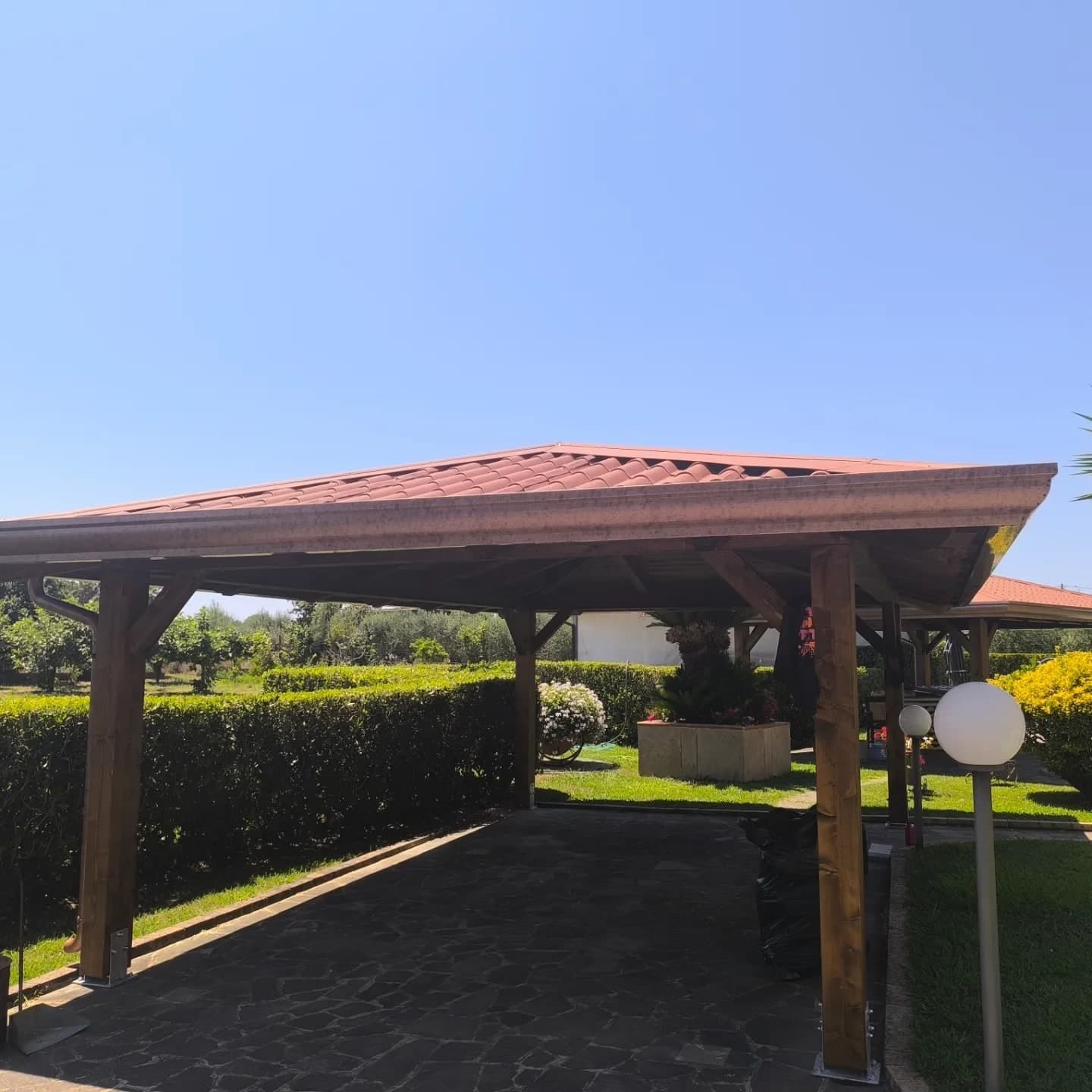Un gazebo in legno con il tetto a padiglione in lastre finto coppo rosse: lungo il bordo la gronda color rame, e un pluviale che scende sul pilastro d’angolo. Sotto, un pavimento in pietra scura.