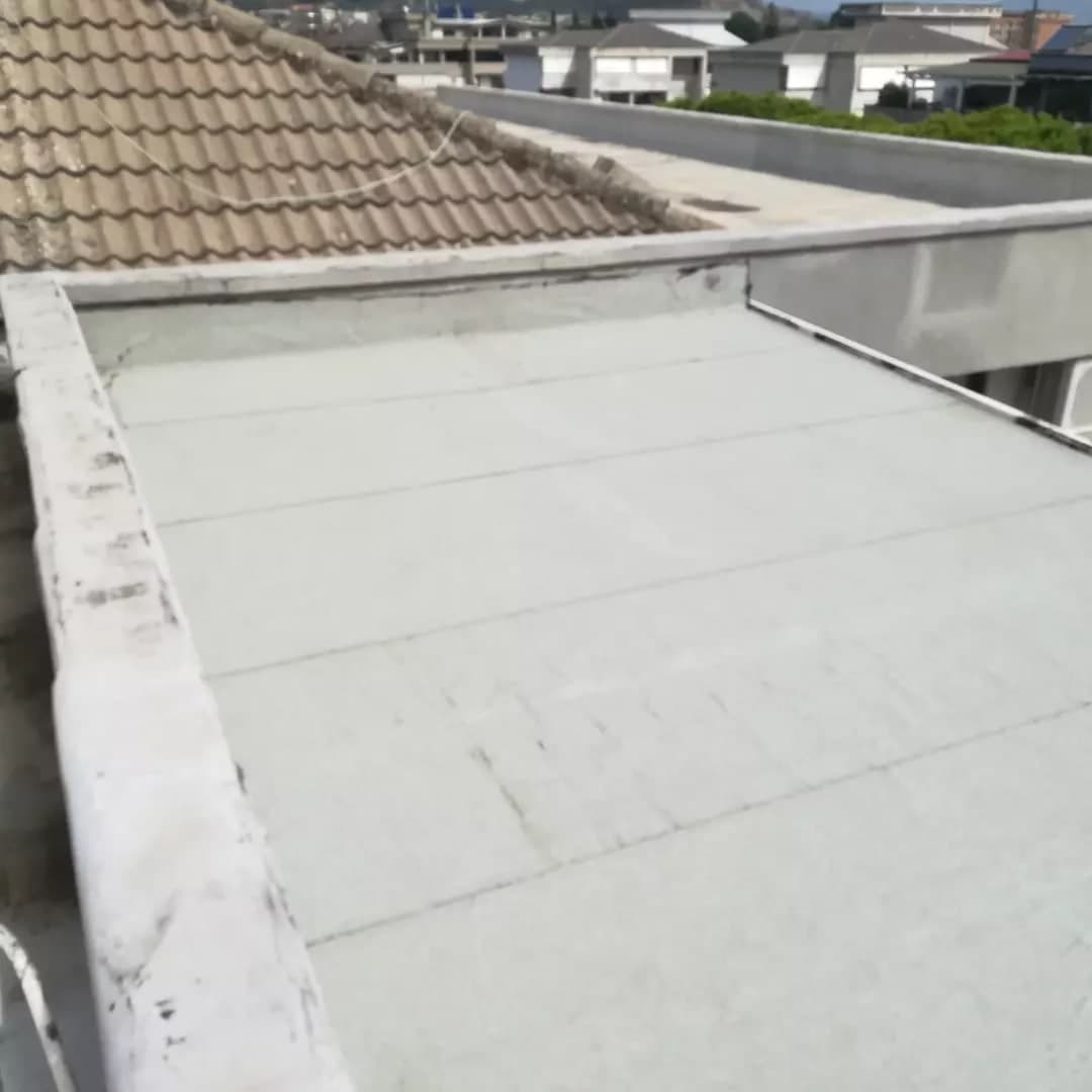 Il tetto della tettoia verso il fondo: la guaina ardesiata chiara a teli lungo il davanti; a sinistra la testa del muro coperta di guaina, in fondo la guaina che risale sul muro fin sotto la copertina, a destra il bordo con la gronda; dietro un tetto in tegole e le case attorno.