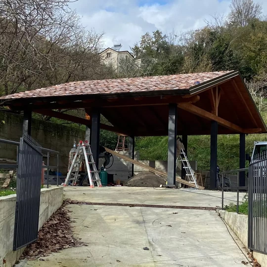 Una tettoia a capanna su un piazzale in fondo a una rampa: il manto in coppi anticati, i pilastri in ferro scuri e, sul timpano, la struttura in legno.