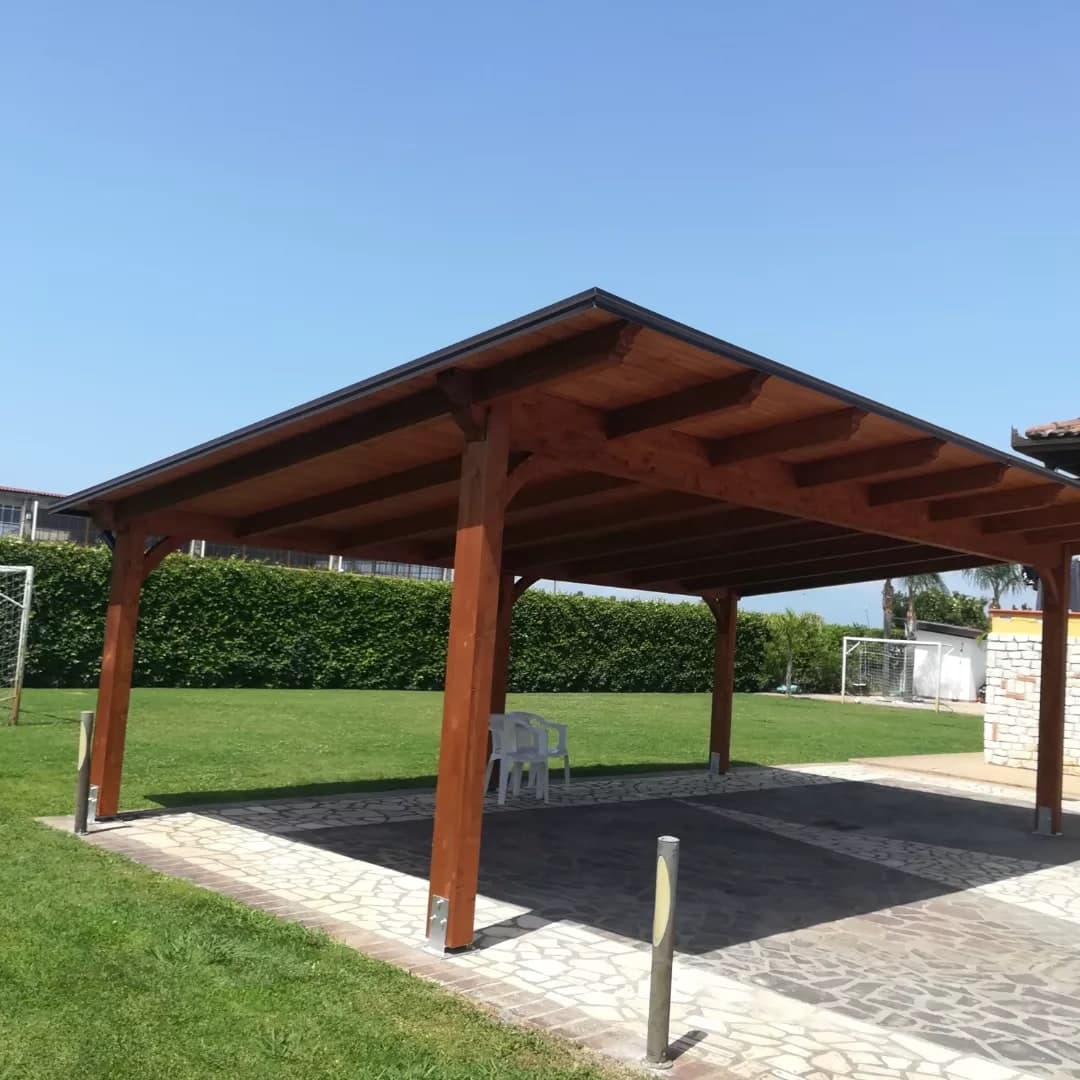 Una tettoia in legno tinto rosso bruno, a capanna, su un pavimento di pietra a opera incerta in mezzo al prato; sotto il timpano le travi e i travetti a vista.