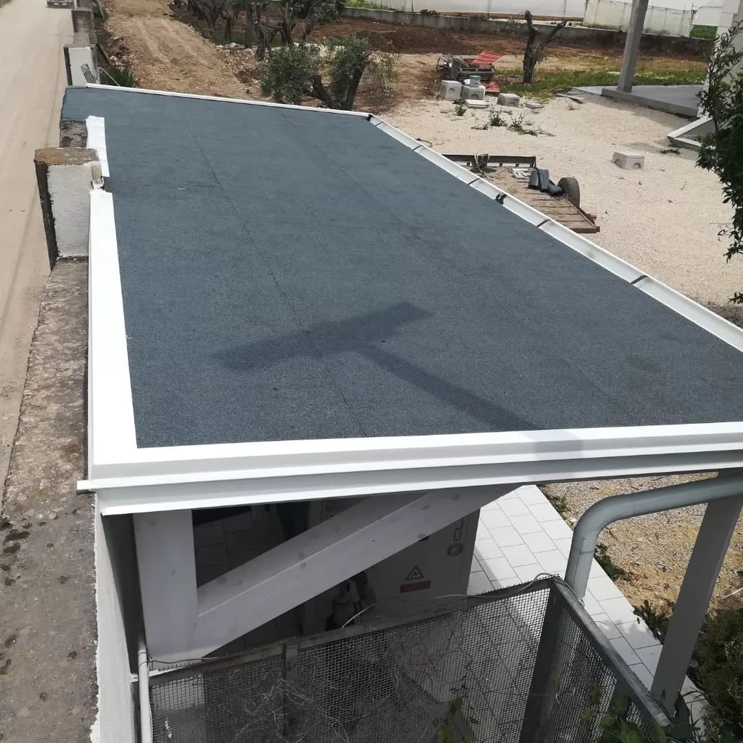Dall’alto, la tettoia lunga e stretta accanto al muro di confine: la guaina ardesiata grigio-blu, la gronda bianca lungo il lato lungo con le staffe scure, la scossalina bianca a gradini sulla testata, il pluviale grigio; sotto, la struttura in legno bianco.
