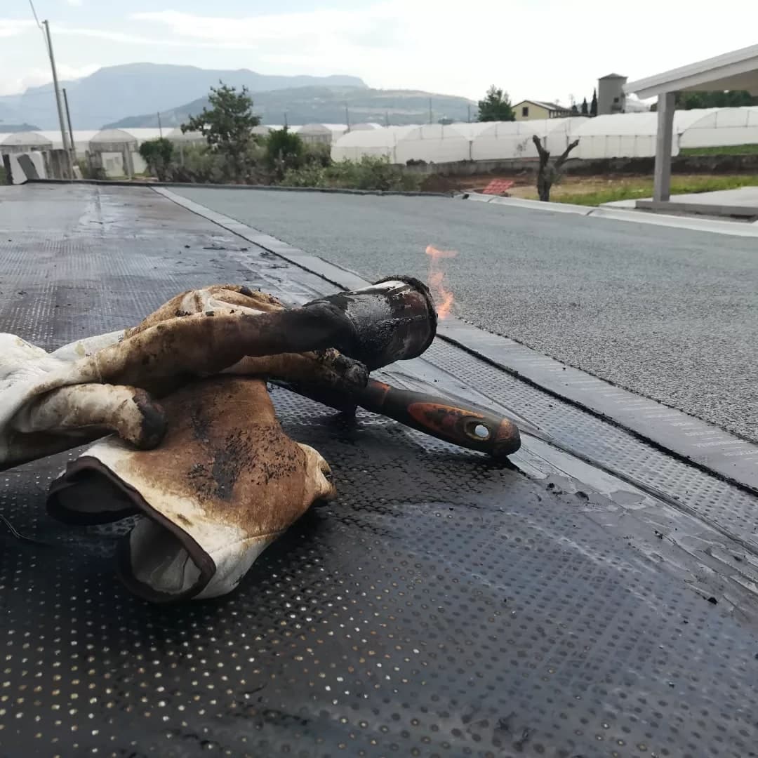 Durante la posa: il primo strato di guaina, nero e stampato a puntini, con i guanti e il cannello acceso posati sopra; più in là la guaina grigia già finita, le serre e i monti.