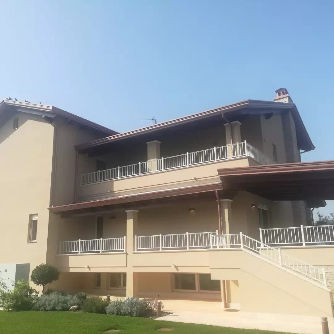 Dal prato, la facciata della villa color crema a gradoni: il seminterrato, il terrazzo del primo piano con la loggia sotto il tetto basso, il terrazzo del secondo piano sotto il tetto grande, la tettoia in legno a destra e la scala esterna; tutti i tetti con lo sporto in legno scuro e la gronda in rame, i pluviali in rame.