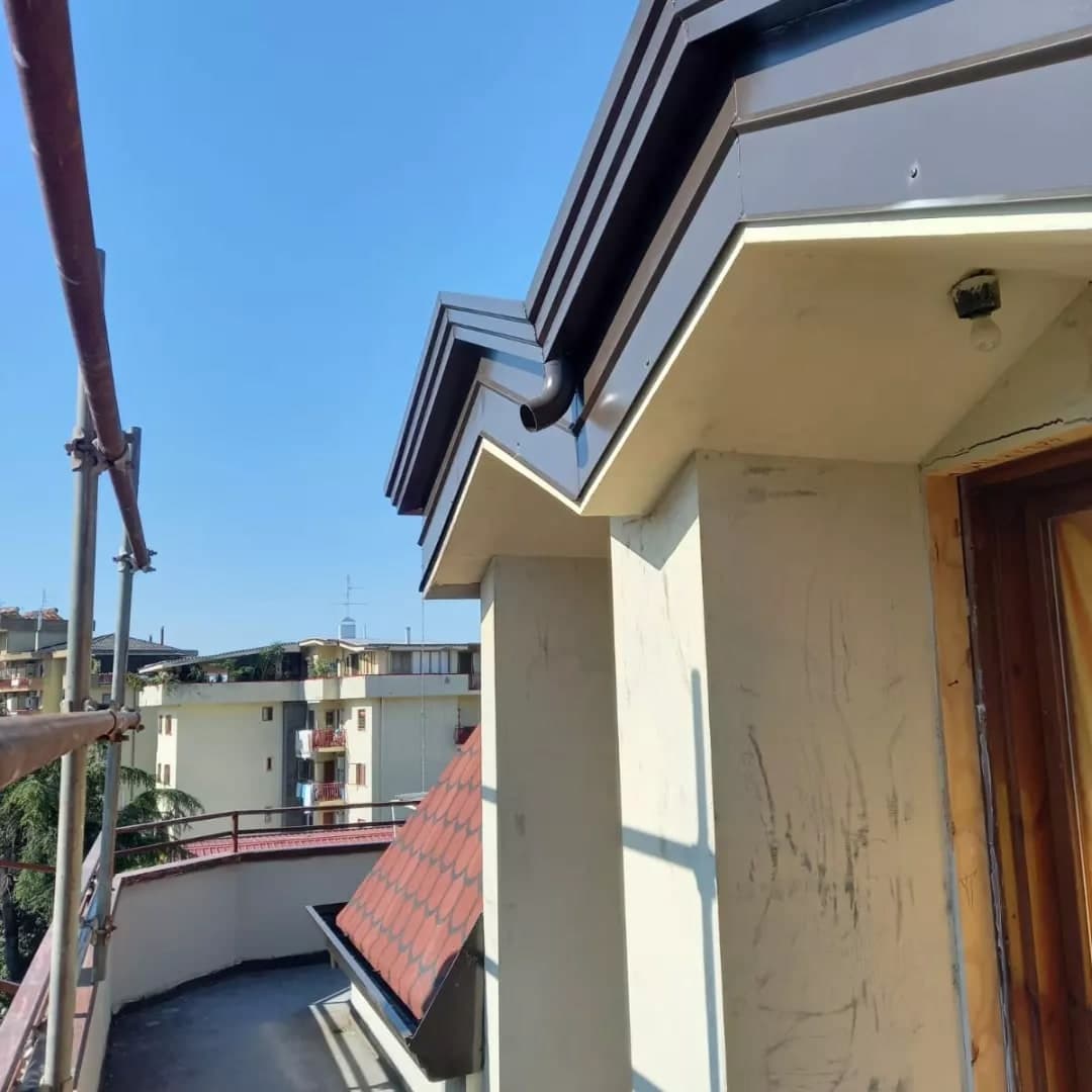 Dal terrazzo dell’attico, guardando in su: la cornice a gradini testa di moro sopra il soffitto chiaro della porta, con il tubo corto che scarica il tetto; più in basso una falda ripida a squame rosse con la sua gronda; attorno il ponteggio e i palazzi.