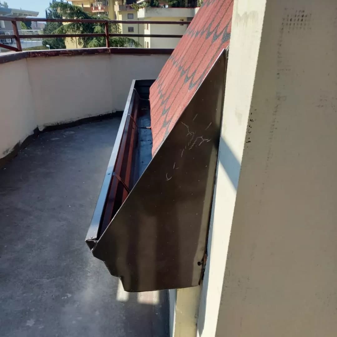 Dall’alto, il capo di una falda sul terrazzo: la gronda quadra testa di moro con le staffe dentro e la grande lamiera scura che chiude la falda e la gronda; a sinistra il parapetto tondo con la ringhiera.