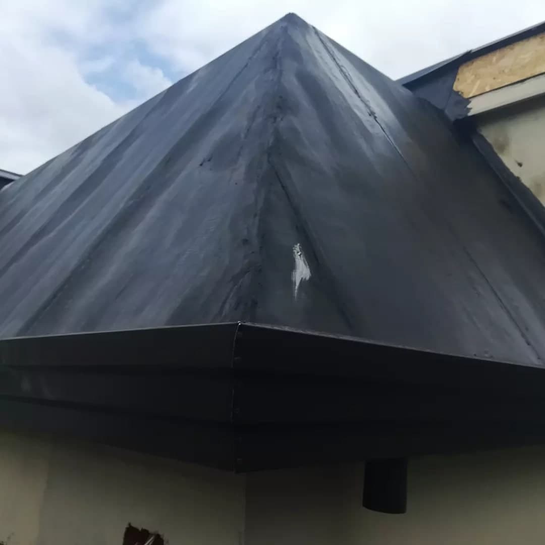 Da sotto, lo spigolo del tetto: il displuvio in guaina nera sale alla punta; sotto, la gronda grande scura a cornice gira l’angolo, con la giunta e la sua colonna di rivetti, e poco dopo scende il pluviale; a destra il muro di un corpo più alto, con i pannelli di legno sopra.