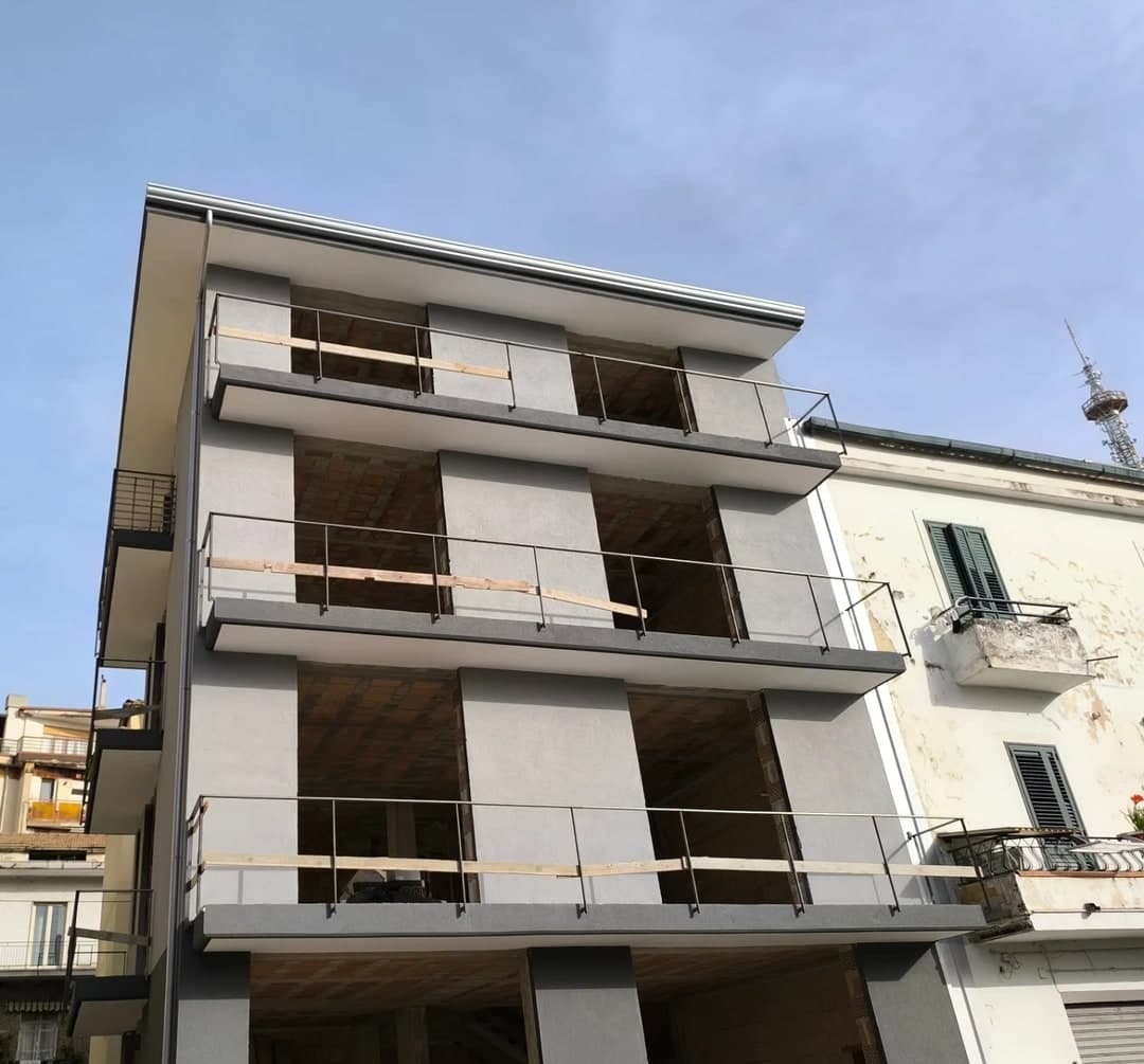 La palazzina grigia dal davanti, da sotto: i balconi con i bordi grigio scuro e le ringhiere, i vani senza infissi; in cima la soletta che sporge, con la gronda grigio chiaro sul bordo, e il pluviale all’angolo.
