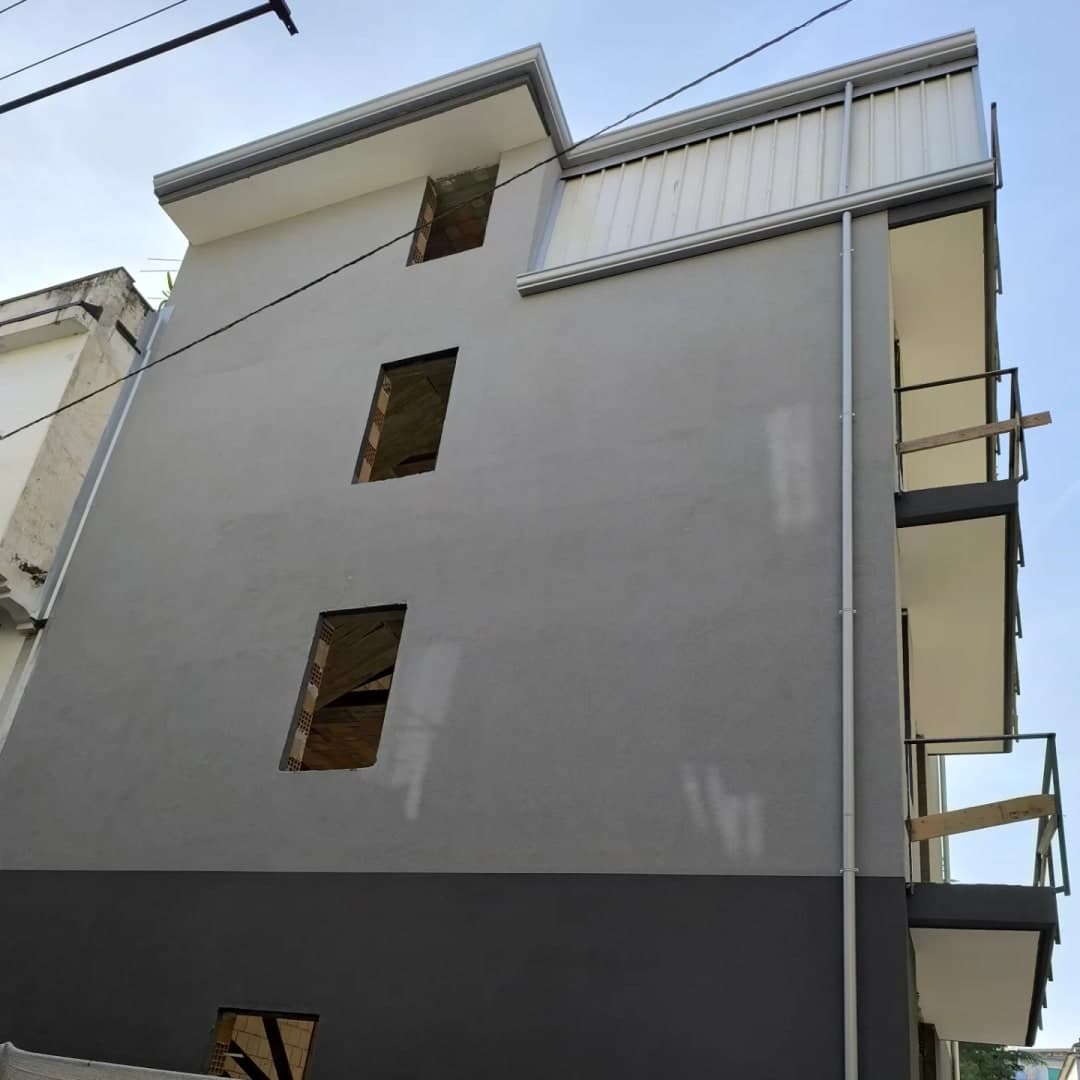 Il fianco grigio della palazzina: in cima la soletta con la gronda grigio chiaro e, accanto, il volume rivestito di lamiera grecata chiara, con la sua cornice; il pluviale scende lungo il fianco fino allo zoccolo scuro.