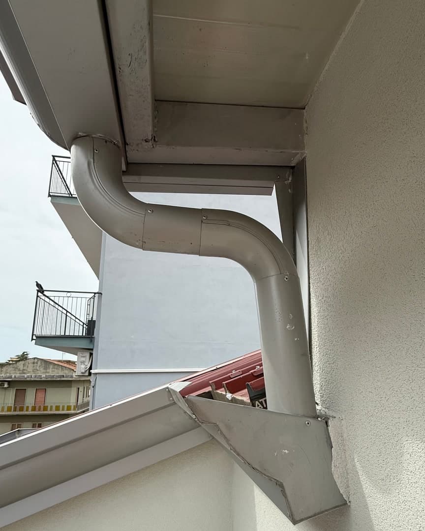 Da sotto: la gronda chiara sotto lo sporto del volume alto e il pluviale che fa una S per tornare al muro, poi scende fino alla tramoggia sopra la gronda di sotto.