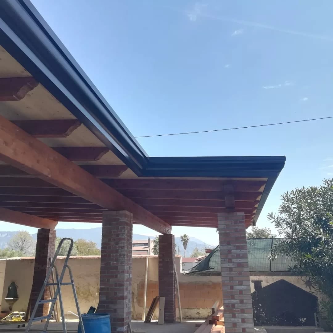 L’angolo di un portico in legno visto da sotto: travi e travetti chiari con il tavolato, pilastri in mattoni faccia a vista, e lungo il bordo la gronda scura che gira l’angolo.