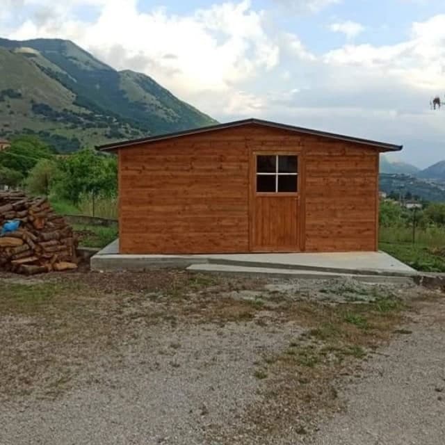 La casetta di fronte, sul piazzale in ghiaia: il timpano con la porta a quattro vetri e il bordo scuro; dietro, i monti.