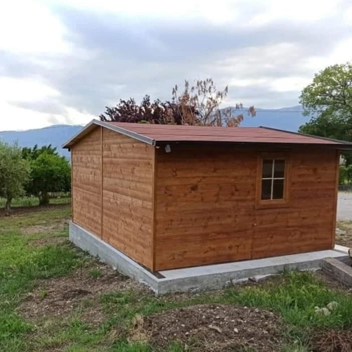 La casetta in legno tinto noce sulla sua platea in calcestruzzo, di tre quarti: il fianco con la finestra, il tetto basso rosso scuro; dietro, ulivi e monti.