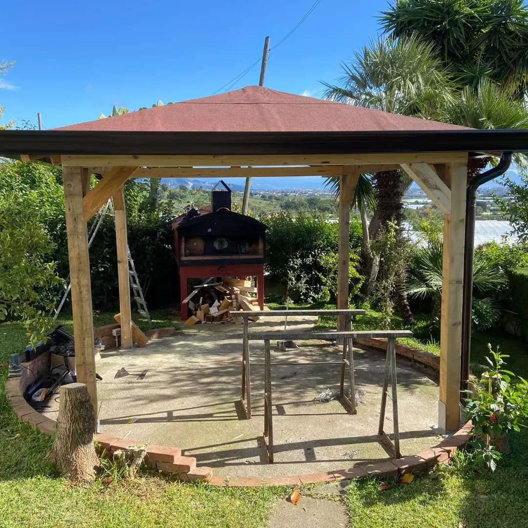 Il gazebo in legno appena finito, sul piazzale chiuso dal cordolo di mattoni: il tetto a padiglione rosso, la gronda nera tutto intorno, il pluviale nero con il collo d’oca sul pilastro di destra; dietro, il forno nella sua casetta rossa e la piana fino ai monti.