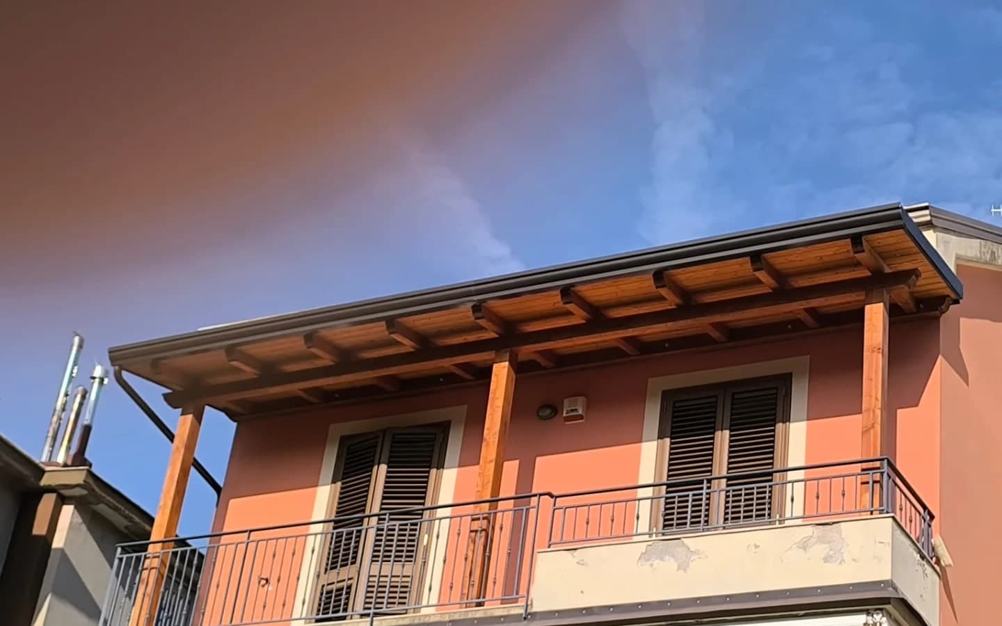 La facciata salmone dell’ultimo piano: il balcone con la ringhiera, i tre pilastri in legno, i travetti a vista e la gronda scura sul bordo della copertura.