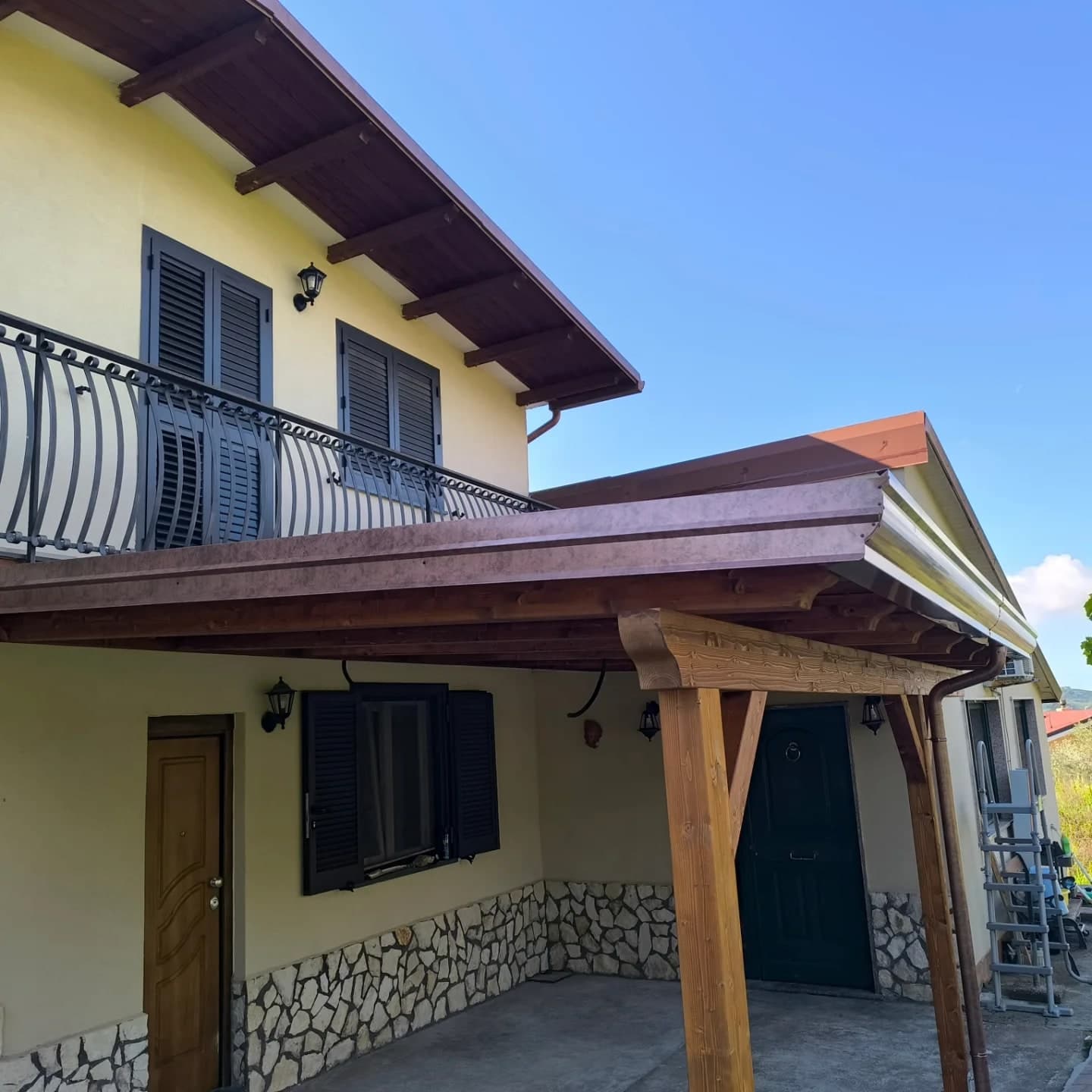 Da sotto, la tettoia in legno davanti alla casa gialla: la gronda a cornice marrone ramato, i due pilastri con le saette, il pluviale; sopra, il balcone con la ringhiera panciuta e il tetto con i travetti a vista.