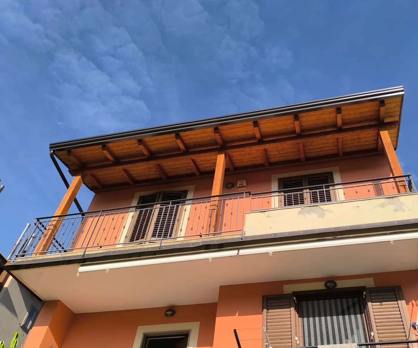 Casa a due piani vista dal basso: sul terrazzo dell’ultimo piano una tettoia in legno con gronda, pluviale e scossaline scure.