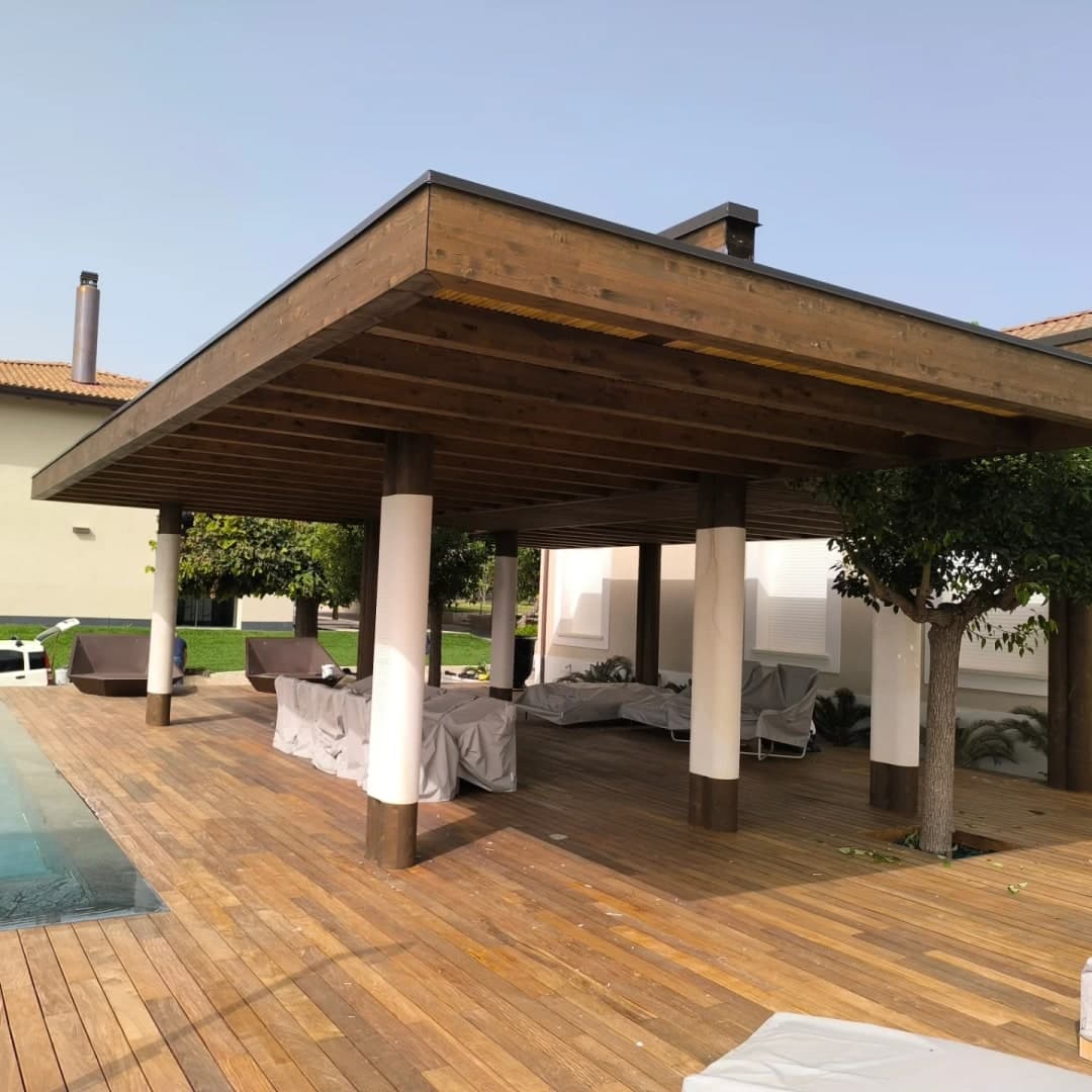 Una grande pergola dal tetto piano su un ponte in legno accanto alla piscina: colonne tonde bianche con la base di legno scuro, travi e travetti in legno, e tutto attorno il bordo in legno con la copertina scura.